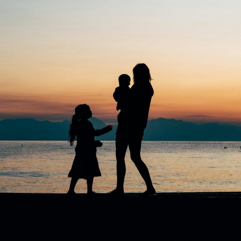 Silhouette einer erwachsenen Person mit zwei Kindern am Seeufer bei Sonnenuntergang.